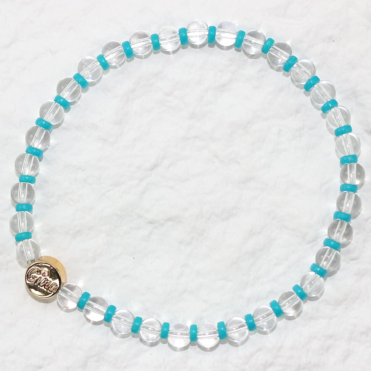A stretch bracelet made of clear crystal quartz beads with vivid aqua-blue spacers and a gold GinaKim logo charm, displayed on a soft white background.