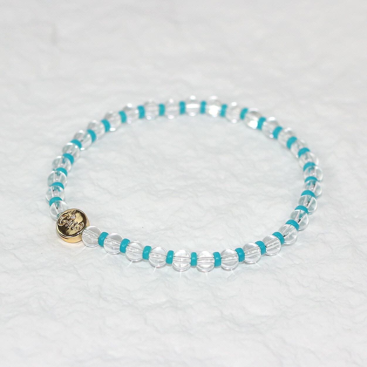 A stretch bracelet made of clear crystal quartz beads with vivid aqua-blue spacers and a gold GinaKim logo charm, displayed on a soft white background.