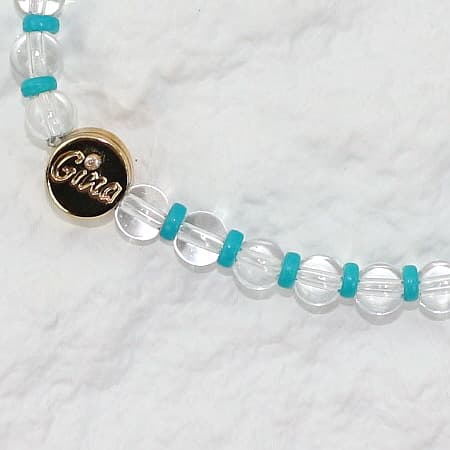 Closeup picture, A stretch bracelet made of clear crystal quartz beads with vivid aqua-blue spacers and a gold GinaKim logo charm, displayed on a soft white background.