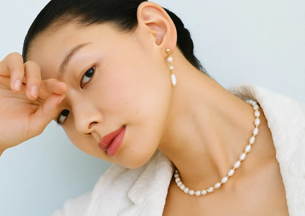 Close-up of an elegant Asian woman wearing a white textured blouse, showcasing a freshwater pearl necklace and gold-accented pearl drop earrings from GinaKim Jewelry