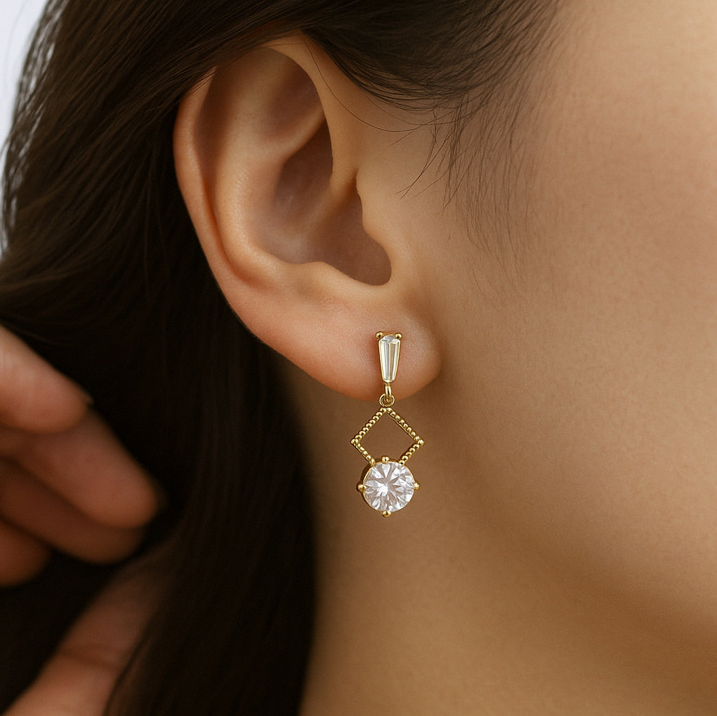A close-up of a woman wearing a gold drop earring featuring a baguette-cut CZ top, an open diamond-shaped frame, and a round brilliant-cut cubic zirconia dangle. Elegant and refined design in warm lighting.