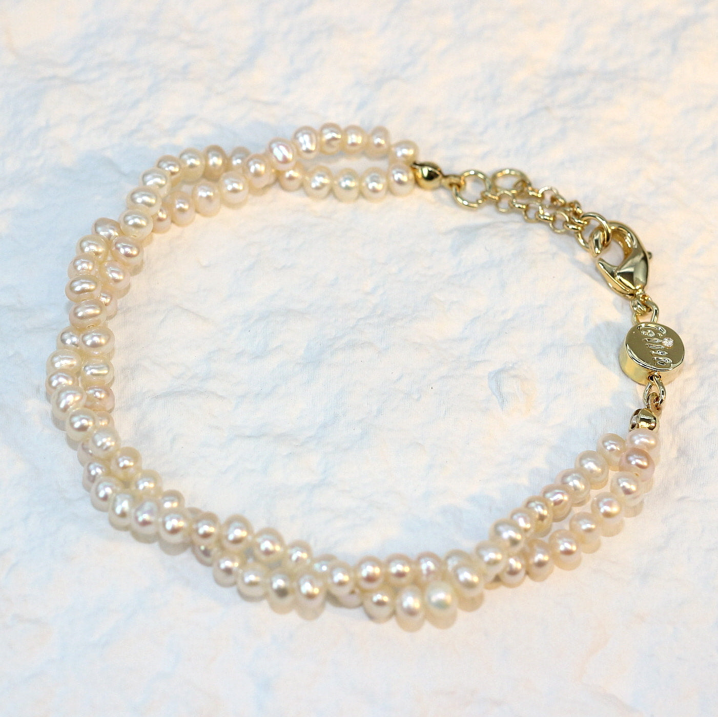 Double Grace Pearl Bracelet displayed flat on a soft white fabric background. The bracelet features two rows of natural freshwater pearls and a gold-plated extender clasp, highlighting its soft luster and layered elegance