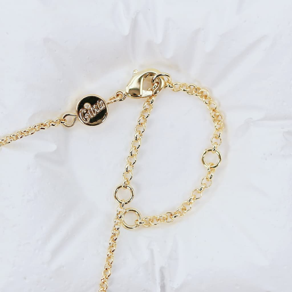 Close-up of a gold-plated necklace showing its lobster clasp and a round logo tag engraved with “Gina,” highlighting the necklace's construction and detail on a white textured background.