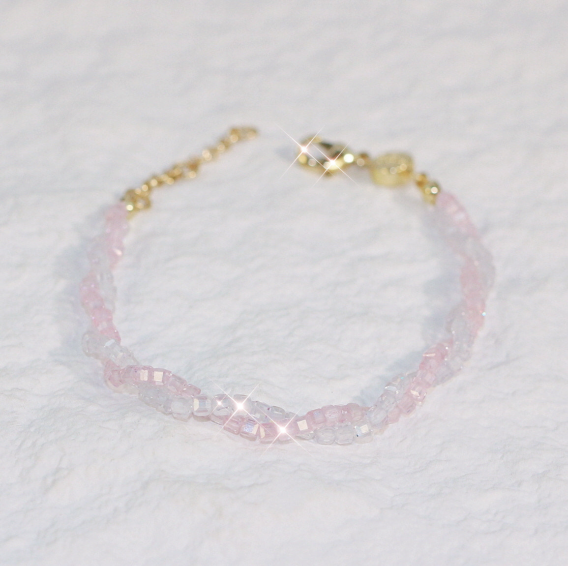 Flat-lay view of the Blush Prism Bracelet showing braided pink and clear cubic zirconia beads with a gold clasp and extender, displayed on a neutral surface.