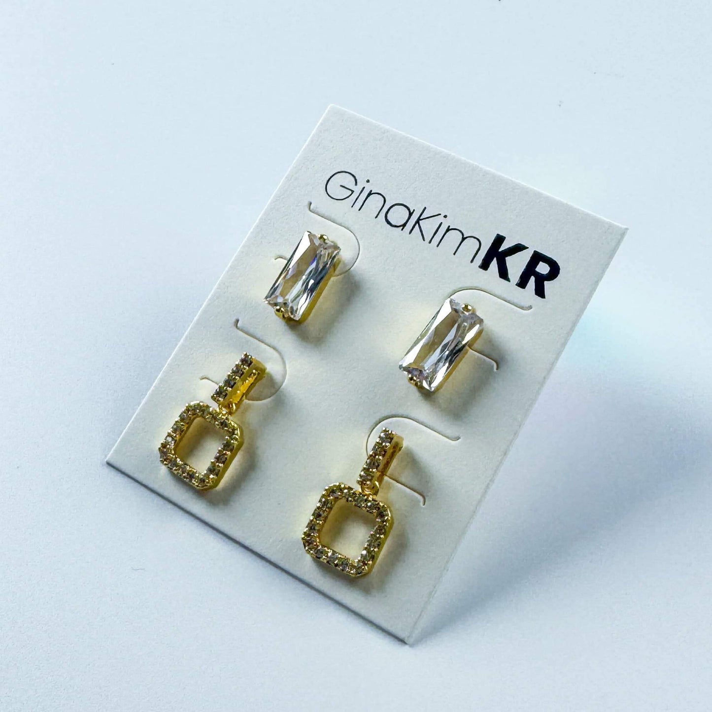 Top view of GinaKimKR Crystal Edge Duo Earrings showing clear crystals and gold pavé drops on white display card