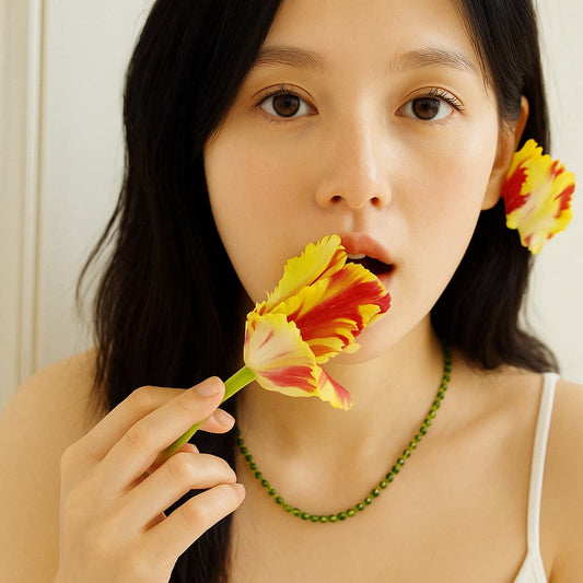 Close-up of a young East Asian woman wearing the Forest Light Necklace by GinaKimKR, holding a red-yellow tulip near her lips. Shot in soft lighting with a natural makeup look.