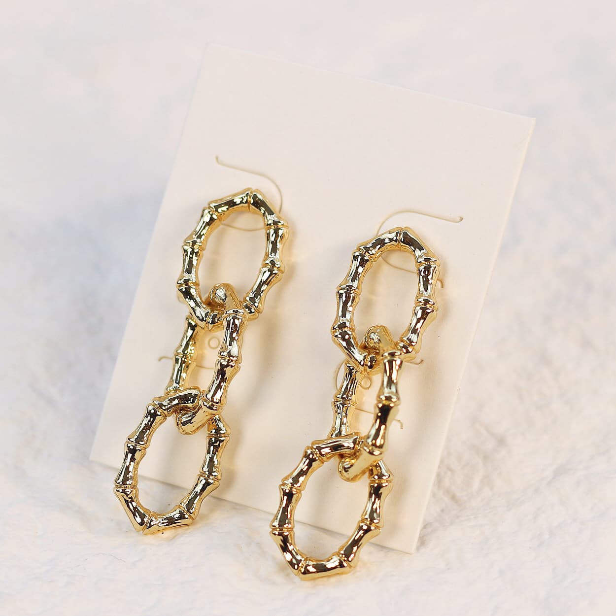 Gold-tone bamboo link drop earrings with three sculpted oval rings and post-back closure, displayed neatly on a cream product card.