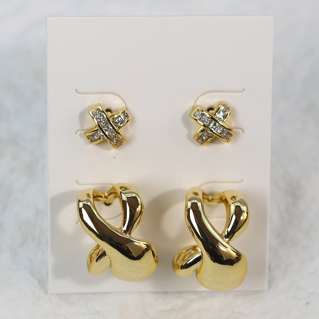 Close-up of gold-tone sculptural earrings and crystal-accented crisscross studs arranged side by side on a white background.