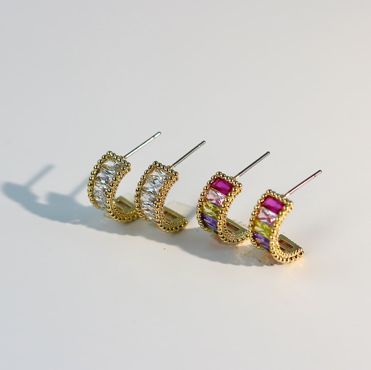 Side shot of Lumière Arc Earring Set highlighting crystal detail—clear CZ baguette pair and multicolor crystal pair on a soft cream background.