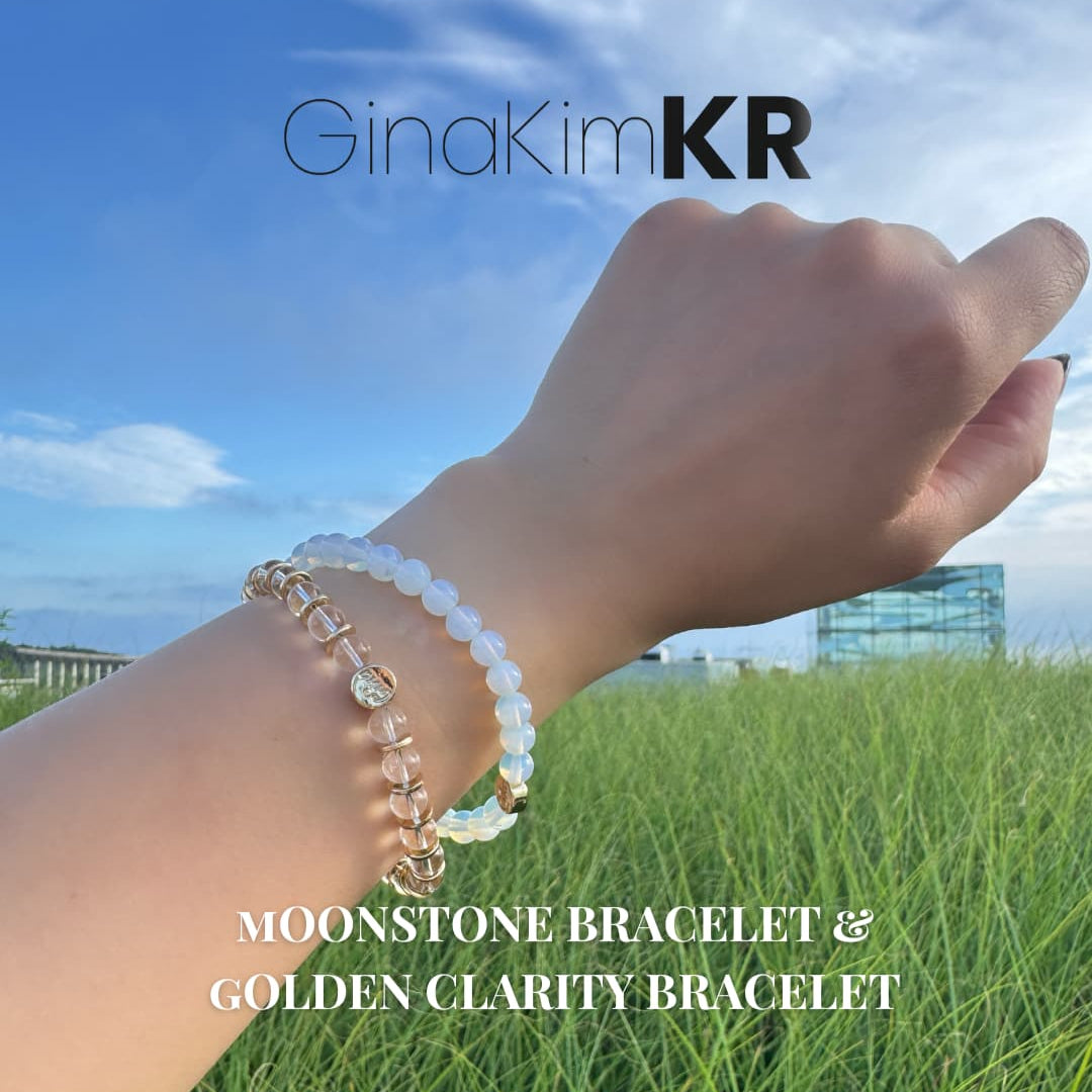 A wrist wearing a stacked combination of the Moonstone Bracelet and Golden Clarity Bracelet from GinaKimKR. Features natural moonstone and crystal quartz beads, styled under natural light with green grass and blue sky in the background.