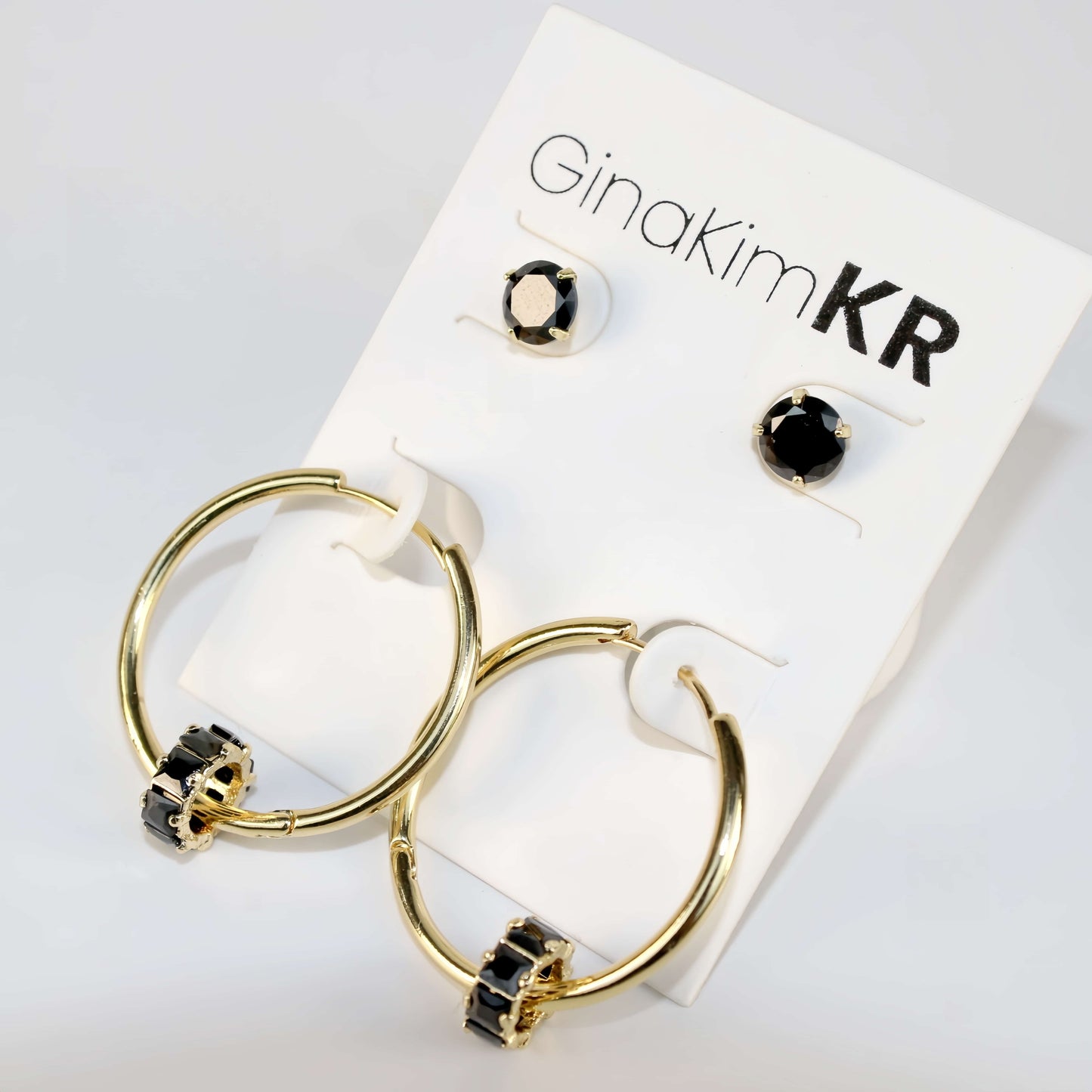 Angled close-up of Noir Balance Earring Set showing gold hoops with jet crystal detail and bold black CZ studs on a cream-toned product card.