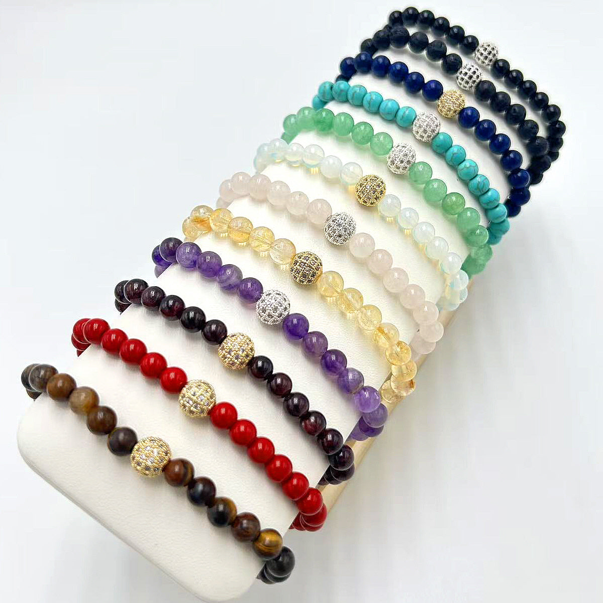12 colorful Basic Fortune Stretch Bracelets from Gina Kim Jewelry displayed on a white stand. Includes natural stones like Tiger’s Eye, Red Agate, Amethyst, Citrine, Rose Quartz, Moonstone, Aventurine, Turquoise, Lapis, Onyx, and more. Elegant semi-precious gemstone bracelets with CZ beads for everyday luck and style.
