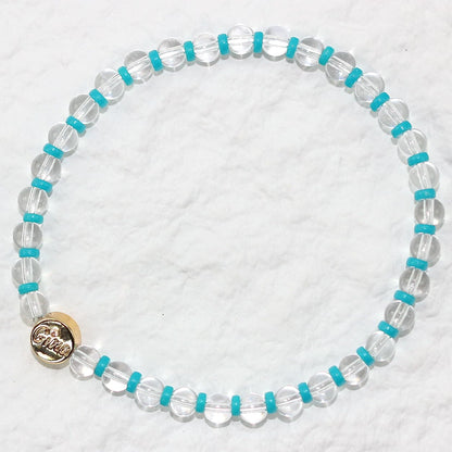 A stretch bracelet made of clear crystal quartz beads with vivid aqua-blue spacers and a gold GinaKim logo charm, displayed on a soft white background.