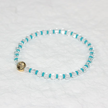 A stretch bracelet made of clear crystal quartz beads with vivid aqua-blue spacers and a gold GinaKim logo charm, displayed on a soft white background.