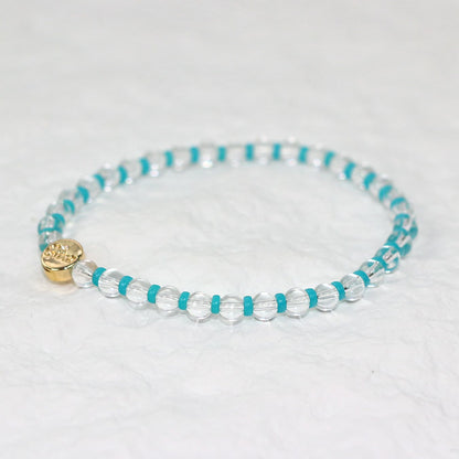 A stretch bracelet made of clear crystal quartz beads with vivid aqua-blue spacers and a gold GinaKim logo charm, displayed on a soft white background.