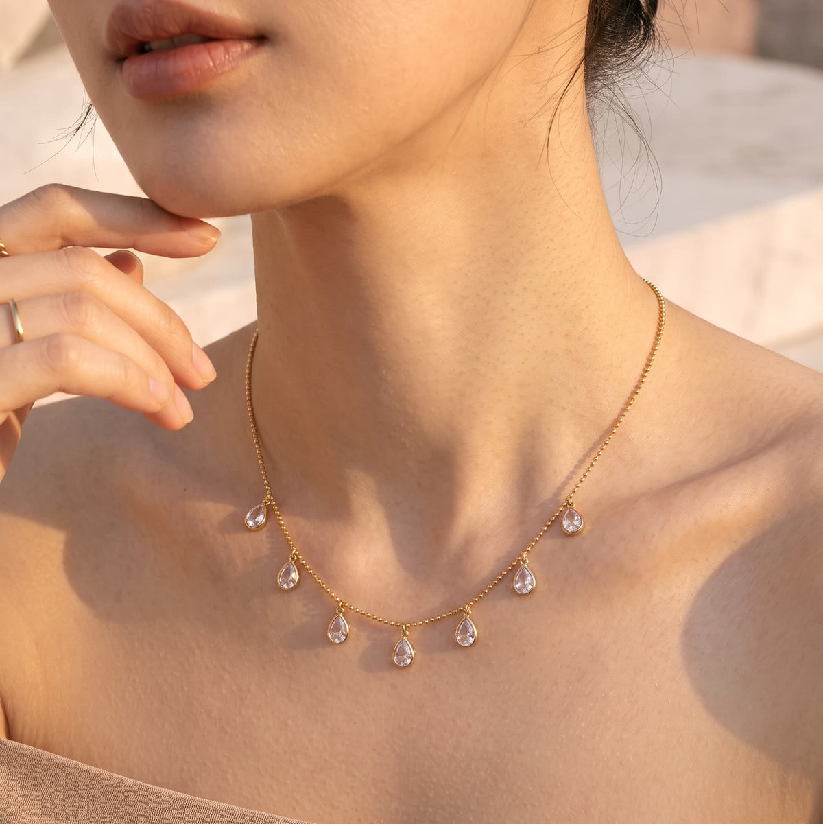 Close-up shot of a woman wearing the Aria Teardrop Station Necklace by GinakimKR in gold. Minimalist daily gold beaded chain necklace with clear Czech glass stones.