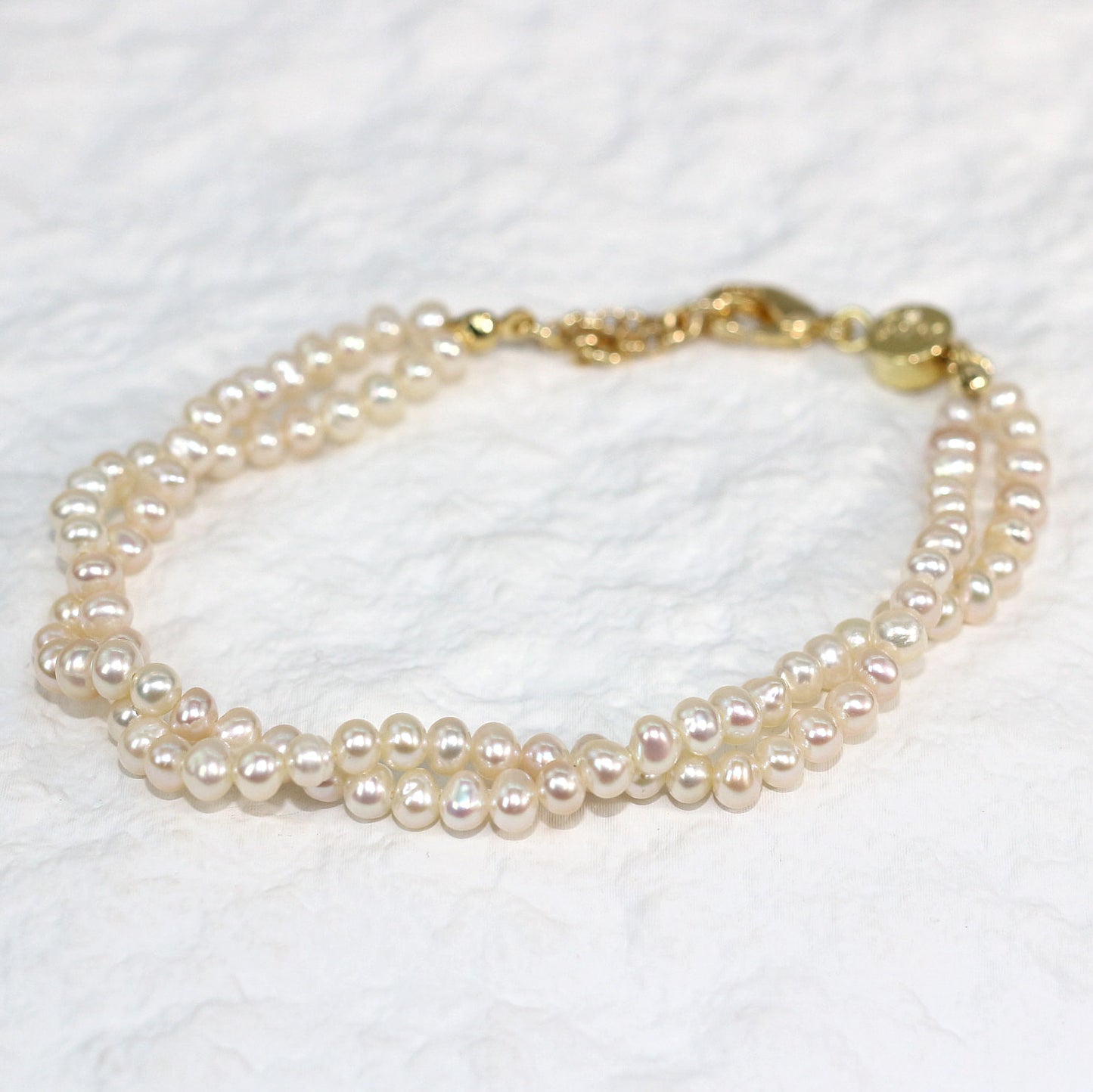 Double Grace Pearl Bracelet displayed flat on a soft white fabric background. The bracelet features two rows of natural freshwater pearls and a gold-plated extender clasp, highlighting its soft luster and layered elegance