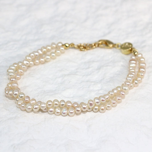 Double Grace Pearl Bracelet displayed flat on a soft white fabric background. The bracelet features two rows of natural freshwater pearls and a gold-plated extender clasp, highlighting its soft luster and layered elegance