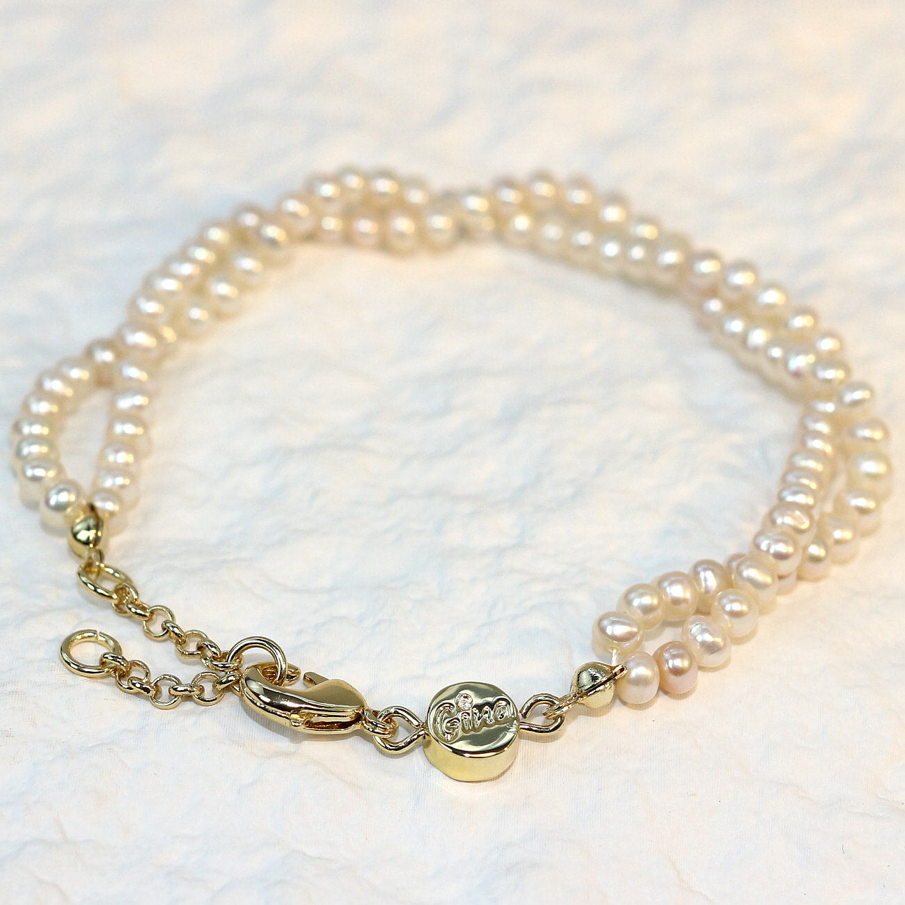 Double Grace Pearl Bracelet displayed flat on a soft white fabric background. The bracelet features two rows of natural freshwater pearls and a gold-plated extender clasp, highlighting its soft luster and layered elegance