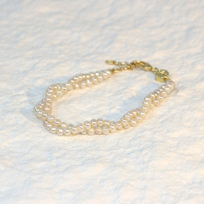 Double Grace Pearl Bracelet displayed flat on a soft white fabric background. The bracelet features two rows of natural freshwater pearls and a gold-plated extender clasp, highlighting its soft luster and layered elegance