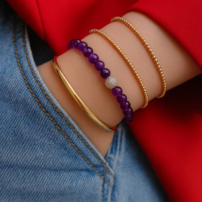 A woman wearing the Amethyst Fortune Bracelet by GinaKimKR, featuring purple amethyst stones, gold detailing, and the signature logo charm. Peaceful and stylish.