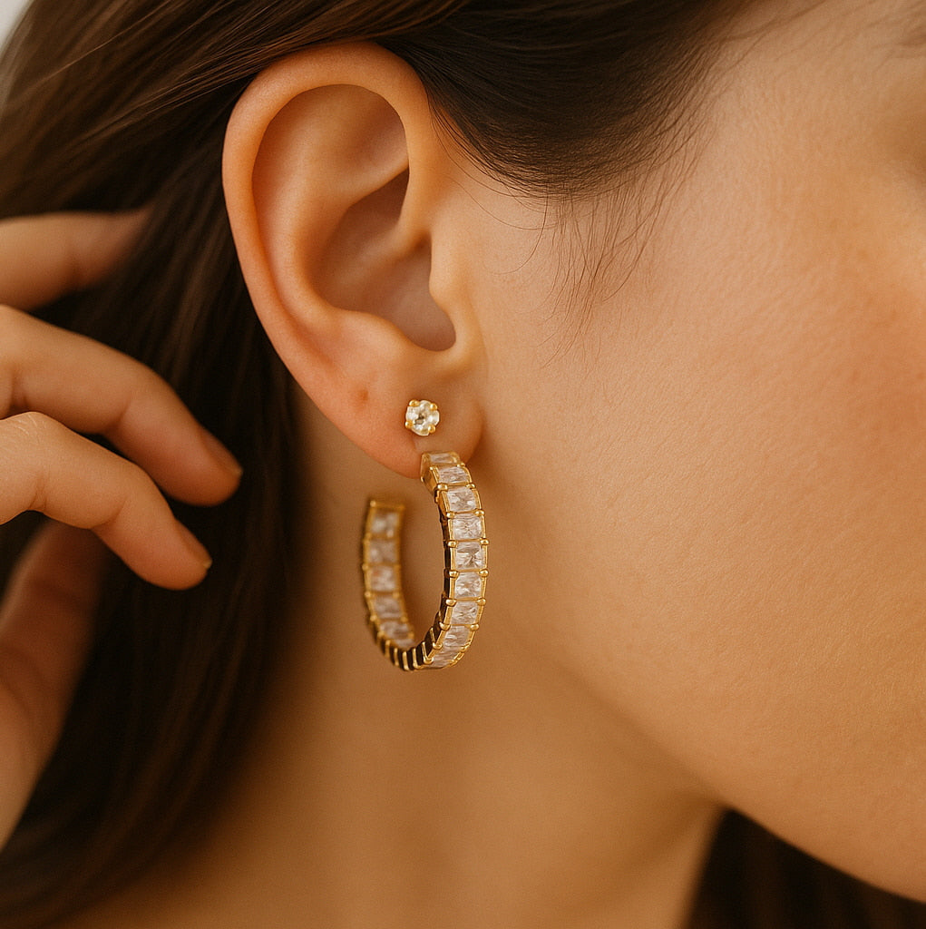 A close-up of a woman's ear wearing a gold open hoop earring with square-cut CZ stones and a small round stud on the upper piercing. The jewelry sparkles softly under warm natural lighting.