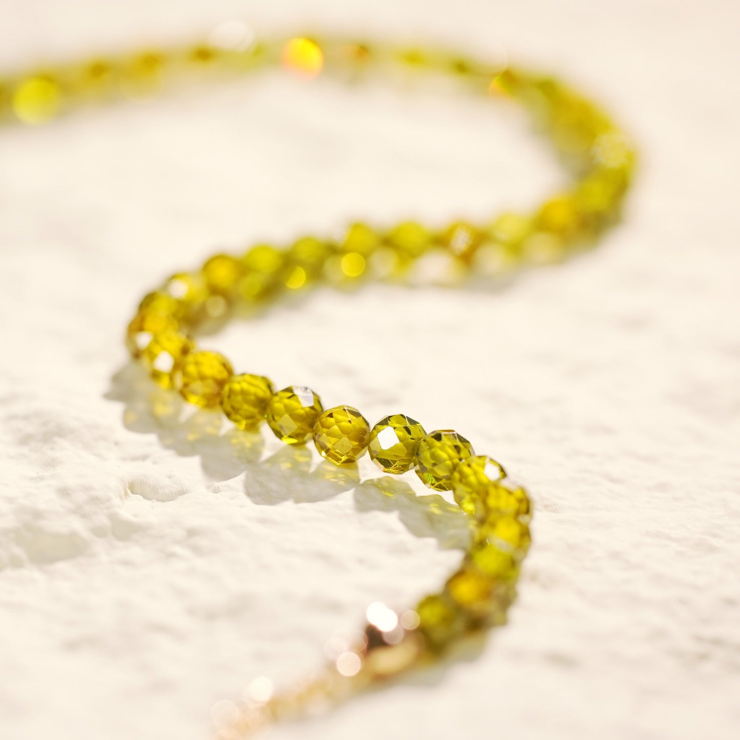Top-down image of the Forest Light Necklace arranged in a gentle S-curve, highlighting the sparkle of forest green CZ beads and handmade gold-toned details on soft fabric.