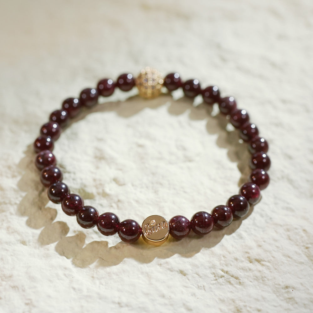 A stretch bracelet with rich red garnet beads, a gold-plated CZ bead, and a Gina Kim logo charm. Bold, elegant, and handmade.