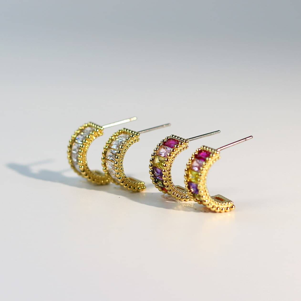 Angled view of Lumière Arc Earring Set showing gold-tone curved earrings, with one pair in clear CZ baguettes and the other in rainbow-colored crystals.