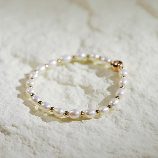 A stretch bracelet crafted with natural freshwater pearls and small gold-plated round beads, featuring a GinaKim logo tag. Displayed on a softly lit, textured background, highlighting the bracelet’s organic elegance and refined Korean design