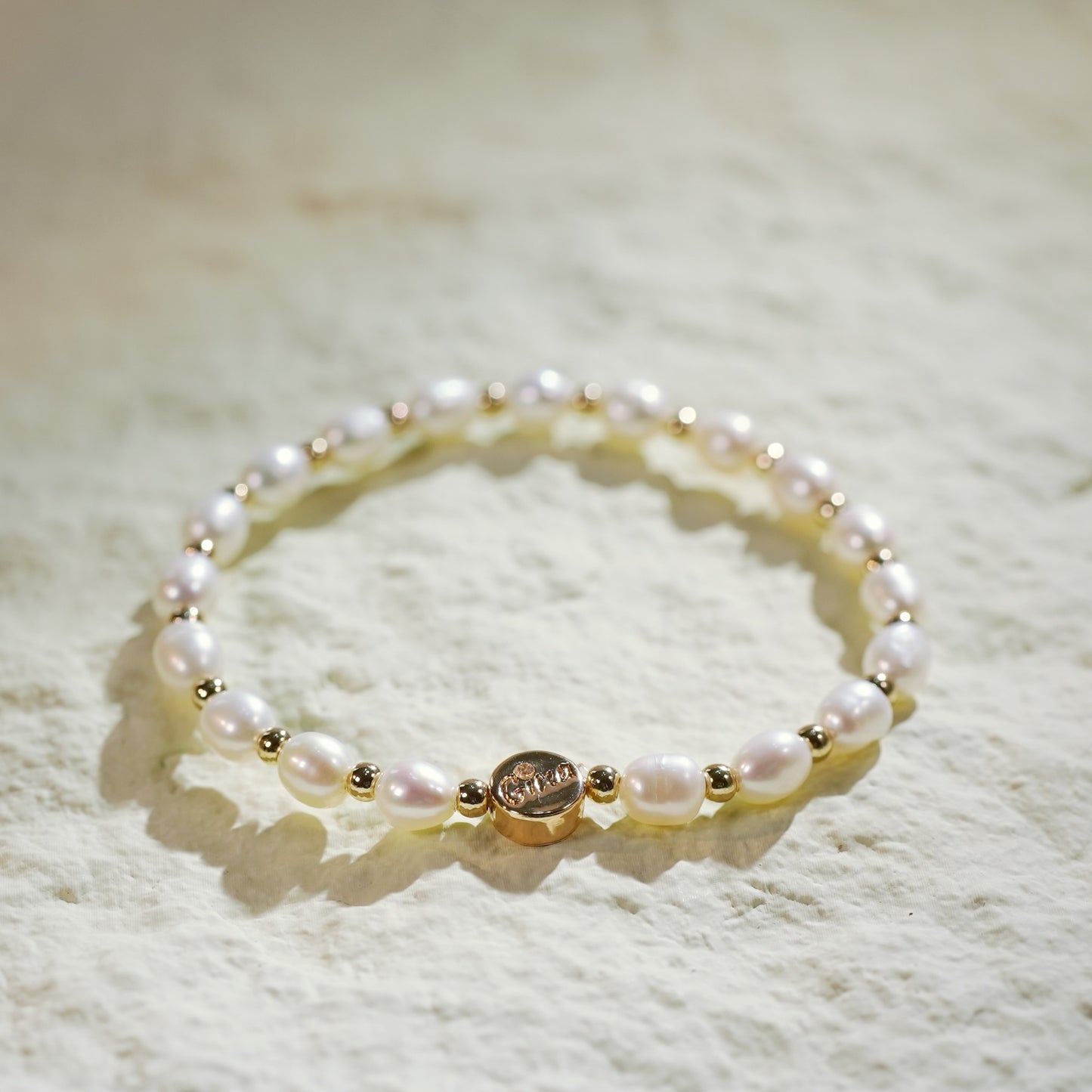 A stretch bracelet crafted with natural freshwater pearls and small gold-plated round beads, featuring a GinaKim logo tag. Displayed on a softly lit, textured background, highlighting the bracelet’s organic elegance and refined Korean design