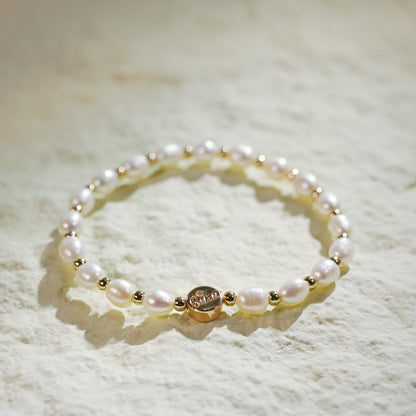 A stretch bracelet crafted with natural freshwater pearls and small gold-plated round beads, featuring a GinaKim logo tag. Displayed on a softly lit, textured background, highlighting the bracelet’s organic elegance and refined Korean design