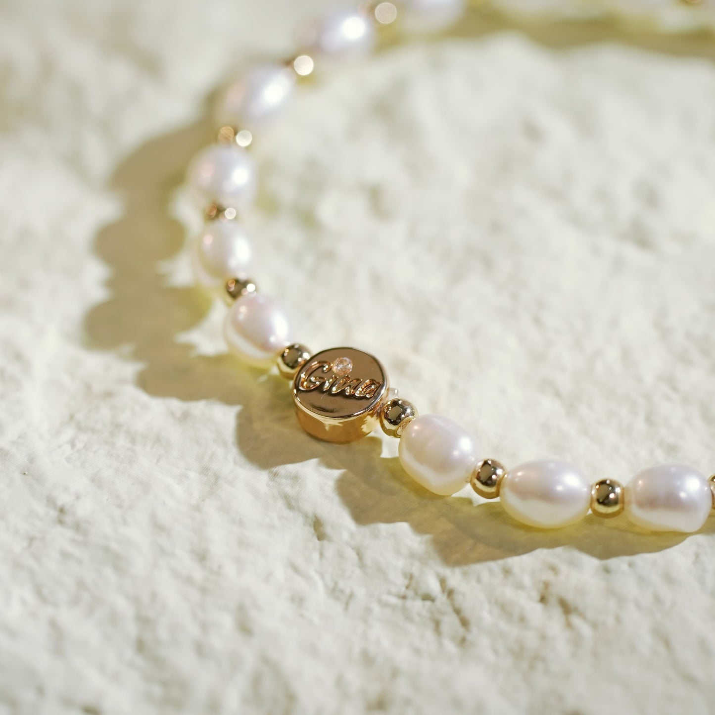 A stretch bracelet crafted with natural freshwater pearls and small gold-plated round beads, featuring a GinaKim logo tag. Displayed on a softly lit, textured background, highlighting the bracelet’s organic elegance and refined Korean design.