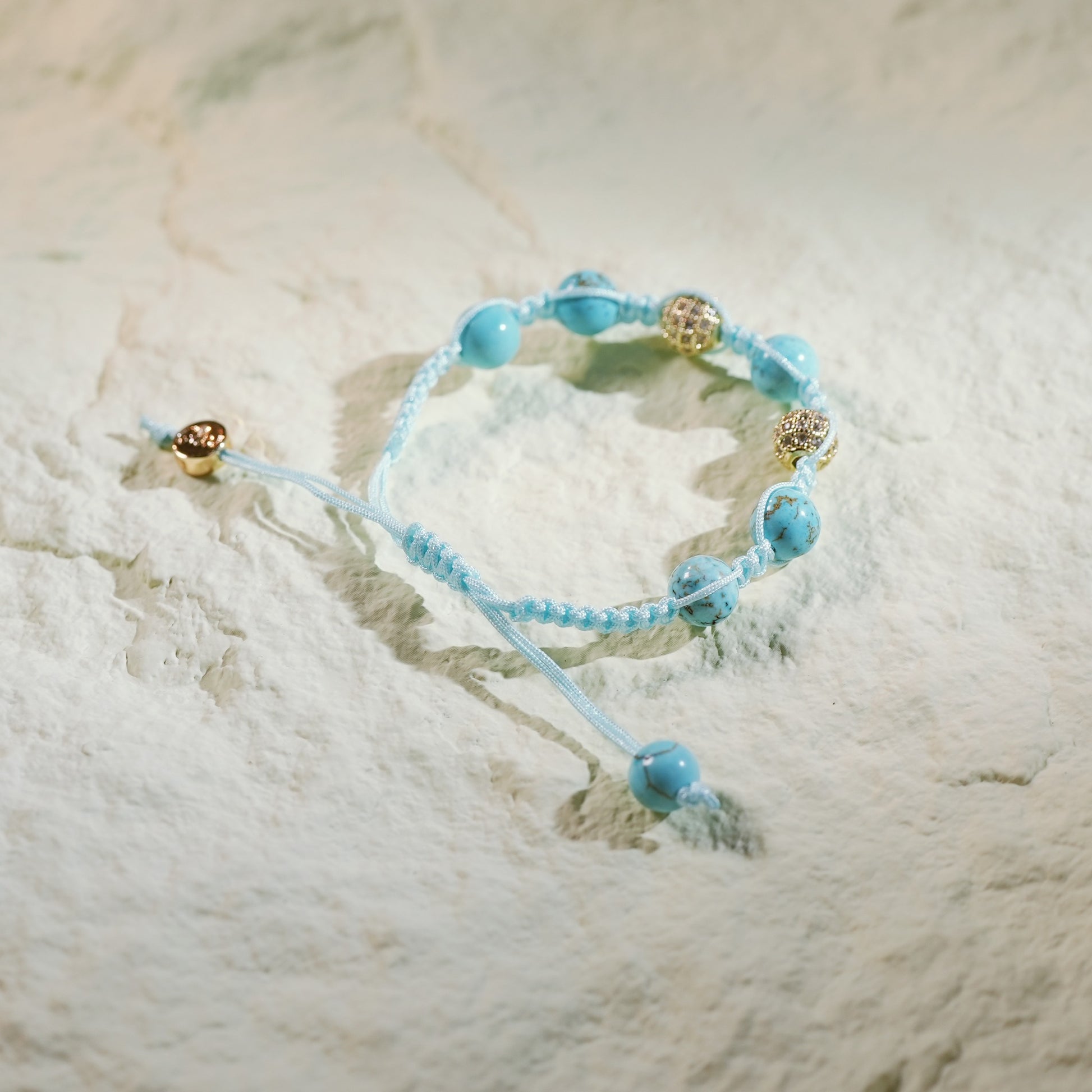 A handmade turquoise macram bracelet featuring natural turquoise beads and gold-plated CZ accents, finished with an adjustable sliding knot and Gina Kim branding. Displayed in a luxury satin-lined gift box